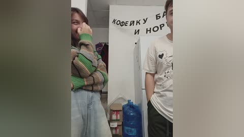 Media: Video of two people in a hallway. One person with brown hair in a green and brown striped sweater is laughing, while the other, wearing a white t-shirt, looks at a sign in Russian.