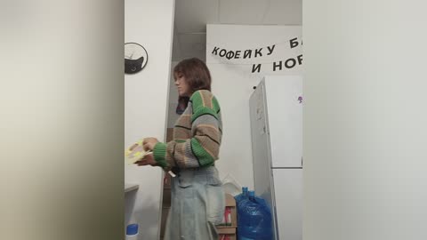 Media: Video of a young woman in a striped sweater, holding a tray, in a white-walled room with Russian signs, a fridge, and blue water bottle.