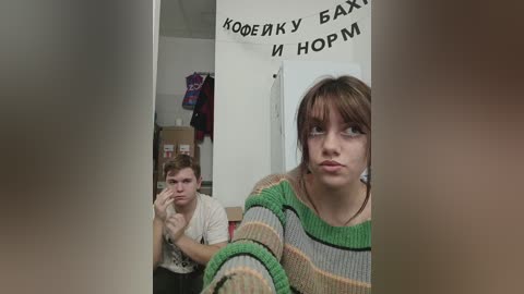 Media: Video of a young woman with brown hair in a striped sweater, standing in a hallway with a surprised expression. A man with short hair and a white t-shirt, holding a phone, is behind her.