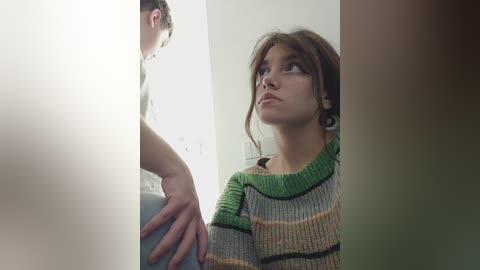 Media: Video of a young woman with light brown hair, wearing a striped sweater, looking upward in contemplation, standing beside a mirror in a bright room.