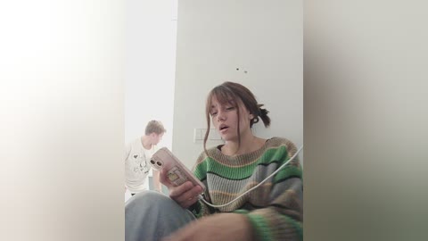 Media: Video of a young woman in a striped sweater, looking at her smartphone, sitting in a white-walled corridor; a man in a white shirt walks by.