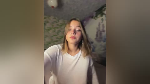 A video of a young woman with light skin and blonde hair, wearing a white long-sleeved shirt, standing in a room with green and white floral wallpaper.