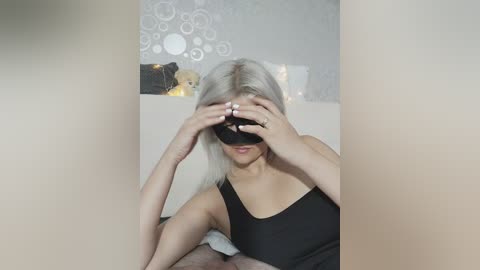 Video of a blonde woman in a black tank top, wearing a black blindfold, sitting on a bed with a white sheet.
