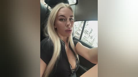 Video of a blonde woman with long hair, wearing a black, unbuttoned shirt, sitting in a car.