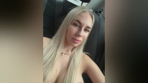 Video of a topless, blonde woman with long hair, fair skin, and a slender physique, seated in a car. She wears a gold necklace and has a serene expression.