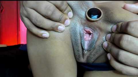 Close-up video of a light-skinned woman's shaved vulva, spread open with her hands. She wears a large, circular, blue gem earring. Background shows a dark, blurry room with red lighting.