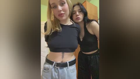 Video of two young women, one blonde and one brunette, wearing black crop tops and high-waisted jeans, standing indoors, smiling, with a blurred background.