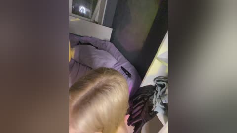 A video of a young blonde-haired boy with a shaved head, wearing a light purple shirt, standing in a dimly lit room with a TV and a window in the background.