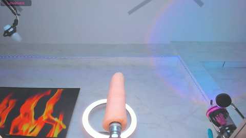 A video of a realistic, pink, phallic-shaped dildo with a white base, placed on a light-colored floor. Background features a blue wall and a black, flame-patterned artwork on the left.