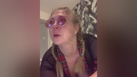 A video of a young woman with blonde braids, wearing oversized, pink-tinted glasses, a black shirt with a red and green plaid scarf, and a floral-patterned curtain in the background.