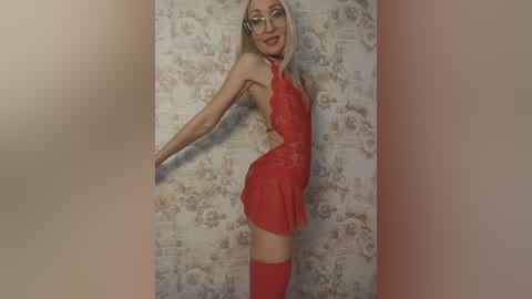 Media: Video of a slender, fair-skinned woman with long blonde hair and glasses, wearing a red lace mini dress, black choker, and red thigh-high stockings, posing against a brick-patterned wall.