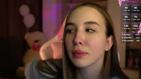 Media: Video of a young woman with fair skin and straight brown hair, wearing a black top, in a dimly lit room. Background includes a teddy bear and a computer screen displaying streaming stats.