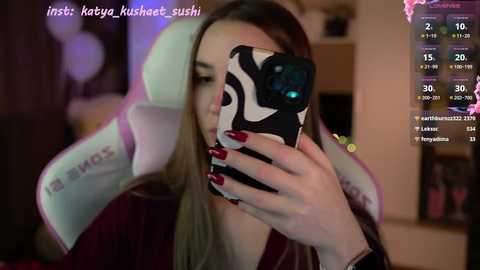 Media: Video of a young woman with long blonde hair, wearing a pink and white gaming chair, taking a selfie in a dimly lit room. She has red nail polish and is holding a smartphone with a black and white case.