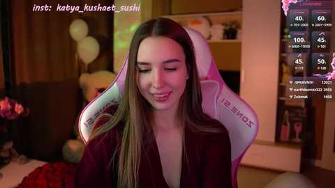 Media: Video of a smiling, light-skinned woman with long brown hair, wearing a maroon shirt, seated in a white and pink gaming chair. Background includes a wooden shelf and dimly lit room.