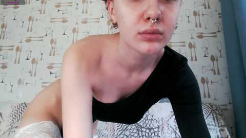 Video of a pale-skinned, androgynous person with short, dark hair, wearing a black one-shoulder top, a septum ring, and a nose piercing. Background features a white wall with a zebra pattern.