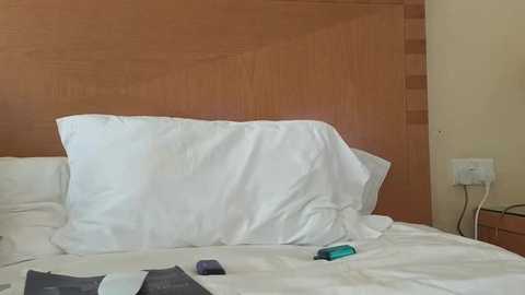 Video of a neatly made bed with white pillows and a wooden headboard, featuring a smartphone, a remote control, and a book on the bedspread. The room has beige walls and an electrical outlet on the right.