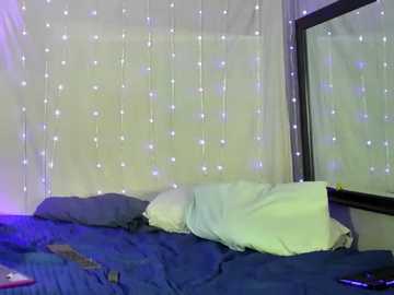 A video of a dimly lit bedroom with a bed covered in blue sheets and a white pillow, a large window with sheer curtains, and fairy lights hanging from the ceiling.