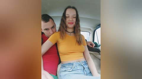 Video of a young couple sitting in a car; man in red shirt, woman in mustard crop top, denim shorts, smiling. Background shows car interior with beige seats.
