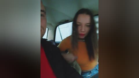 A blurry video captures a young woman with long brown hair, wearing a tight orange crop top, engaging in a sexual act with a man inside a car.