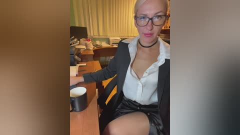 A video of a woman in a black suit jacket and white blouse, glasses, and black choker, sitting at a wooden desk with a coffee mug, in a cluttered office.