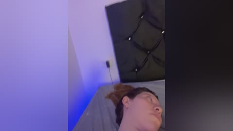 Video of a woman lying on a bed with a dark headboard, eyes closed, wearing a gray shirt, in a dimly lit room with blue lighting.