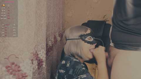 A video depicts a young woman with blonde hair, wearing a black lace mask and blue kimono, performing oral sex on a standing man in a dimly lit, worn-out room.