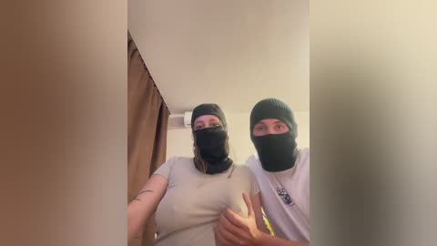 Video of two women, both wearing black balaclavas, light-colored t-shirts, and jeans. They are indoors, standing in a dimly lit hallway, with one woman's hand on the other's shoulder.