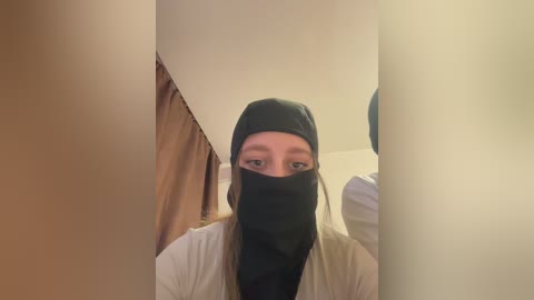 A video of a person wearing a black face mask, beige jacket, and brown hair, standing in a dimly lit room with beige walls and a brown curtain.
