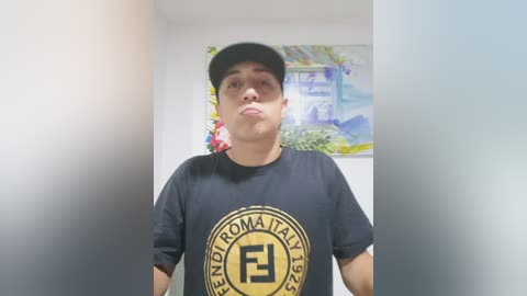 Video of a young man with light brown skin and short black hair, wearing a black baseball cap, T-shirt with Italian text, and making a kissy face in a brightly lit room with floral and mountain art on the wall.