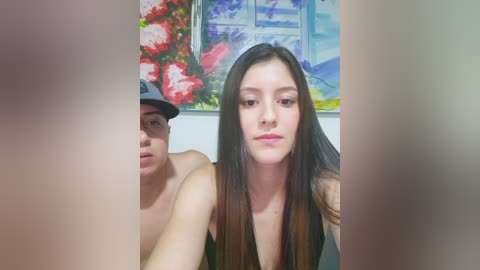 Video of two people: a shirtless man with a cap, and a woman with long brown hair, both indoors with a colorful abstract painting in the background.