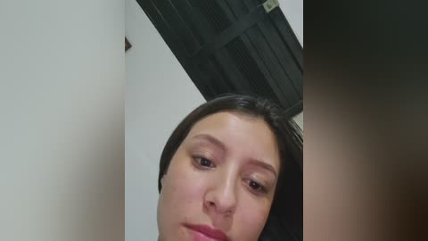 Video of a young woman with pale skin and straight, dark brown hair, looking down at the camera. She has a neutral expression. Background includes a white wall and a black door.