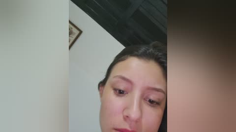 A close-up video of a young woman with fair skin and dark hair, wearing minimal makeup, captured from a low angle. The background features a white wall and a wooden ceiling.