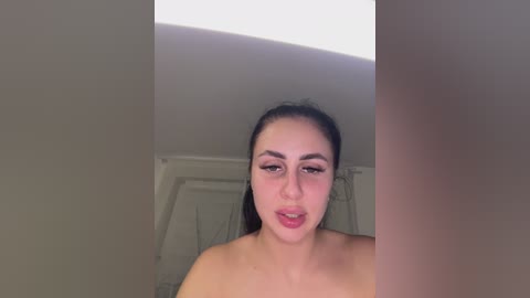 A video of a young woman with light skin and dark hair, captured from a slightly low angle, showing her bare shoulders and upper chest.