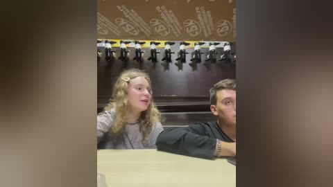 Media: Video of two young people, a girl with curly blonde hair and a boy with short brown hair, seated at a wooden table in a dimly lit room with a large, brown, patterned ceiling.