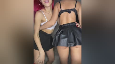 Video of two women with fair skin, one with pink hair, wearing lingerie and short black leather skirt, standing close together against a beige background.
