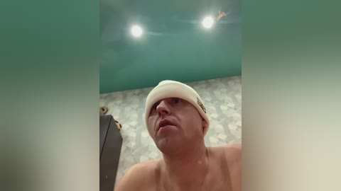 Video of a shirtless, pale-skinned man with a white beanie, looking up, in a room with teal walls and a patterned floor.