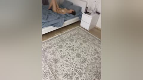 Media: A blurry video of a bedroom with a person lying on a bed, a white nightstand, and a detailed, ornate rug on the floor.