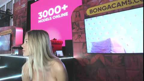 Media: Video of a topless blonde woman with long hair, seated in a room with a pink wall and a large screen showing a bed, surrounded by models online.