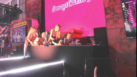 Media: Video of a lively game show set, featuring a pink backdrop, a large \"Bingo\" sign, and four contestants in casual attire, including a blonde woman. Arcade games and monitors are visible in the background.