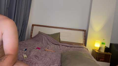 Media: Video of a nude person with light skin, sitting on a bed with a purple quilt, a wooden headboard, and a bedside table with a green lamp.