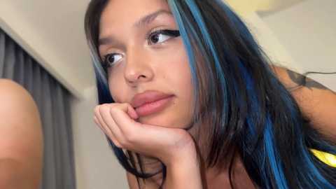 Media: Video of a young woman with long, black hair with blue highlights, light brown skin, and dark eyes, resting her chin on her hand, wearing a yellow top.