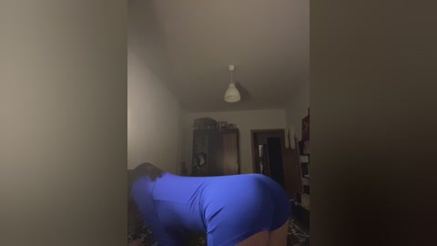 Video of a person in blue pajamas crawling on all fours in a dimly lit room with a white ceiling light and various furniture in the background.
