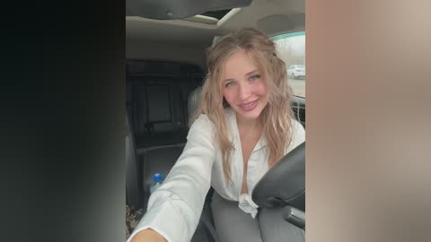 Video of a smiling, light-skinned woman with long, wavy blonde hair, wearing a white blouse and gray pants, driving a car with a gray interior.