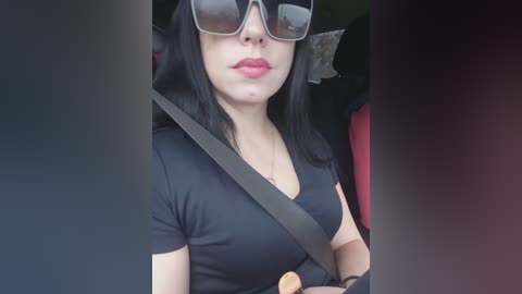 Media: A video of a fair-skinned woman with long black hair, wearing large black sunglasses, a black V-neck top, and a seatbelt, seated in a car.