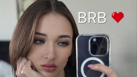 Media: Video of a fair-skinned woman with straight, shoulder-length brown hair, blue eyes, and light makeup, taking a selfie with a black iPhone in a dimly lit room. \"BBR\" logo with a red heart symbol is overlaid.