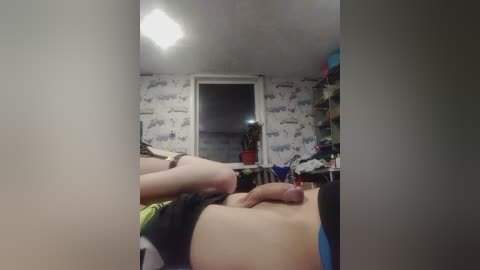 Video of a messy, cluttered bedroom with white wallpaper featuring cartoon patterns. A person with fair skin is lying on a bed, partially visible, with a prominent erect penis. The room is dimly lit, with a bright ceiling light casting shadows.