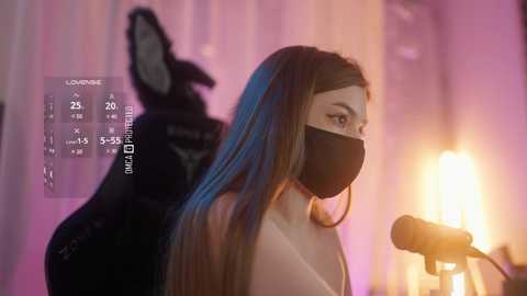 Video of a woman with long, straight brown hair, wearing a black face mask, speaking into a microphone, illuminated by warm, ambient light, with a shadowy figure behind her.
