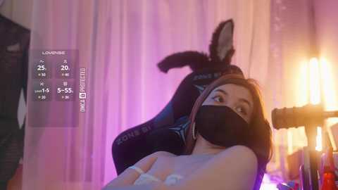 A video featuring a woman in a black face mask, wearing a black hoodie with a white logo, lying on a bed with a black and white striped pillow. The background includes a large lamp and sheer curtains.