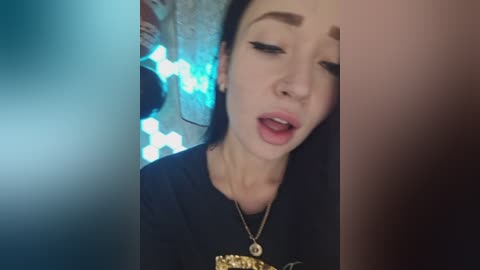 Video of a young woman with pale skin and long dark hair, wearing black clothing and a gold necklace, capturing a moment with a sensual expression. Background features blurred, blue lights and a brick wall.