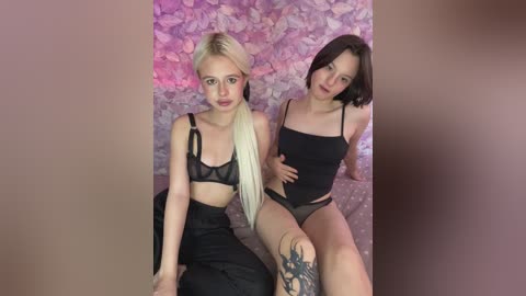 Video of two young women with fair skin and light makeup; one with platinum blonde hair in a high ponytail and black lingerie, the other with dark brown hair in a bob and black lingerie. They sit against a textured, pink floral background.
